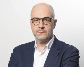 Ein Mann mit Brille und kurzem, grauem Bart trägt einen dunkelblauen Anzug und ein weißes Hemd. Er steht mit verschränkten Armen vor einem hellen Hintergrund.