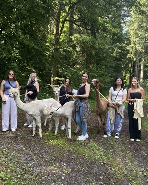 Eine Gruppe von Frauen steht in einem Wald, umgeben von mehreren Alpakas und einem Lama.