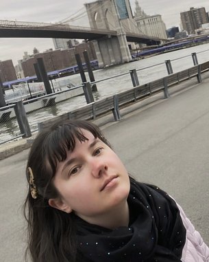 Eine Person mit langen, dunklen Haaren und einem schwarzen Schal steht am Wasser, mit der Brooklyn Bridge und der Skyline im Hintergrund.