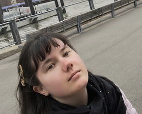 Eine Person mit langen, dunklen Haaren und einem schwarzen Schal steht am Wasser, mit der Brooklyn Bridge und der Skyline im Hintergrund.