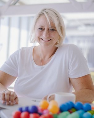 Eine lächelnde Frau mit blonden Haaren sitzt an einem Tisch mit einem Laptop und einer Tasse. Vor ihr liegt eine bunte Ansammlung von kleinen Kugeln.