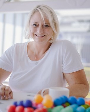 Eine lächelnde Frau mit blonden Haaren sitzt an einem Tisch mit einem Laptop und einer Tasse. Vor ihr liegt eine bunte Ansammlung von kleinen Kugeln.