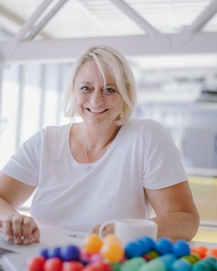 Eine lächelnde Frau mit blonden Haaren sitzt an einem Tisch mit einem Laptop und einer Tasse. Vor ihr liegt eine bunte Ansammlung von kleinen Kugeln.