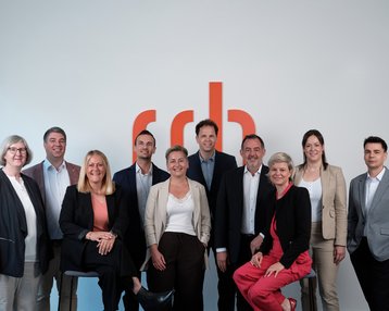Eine Gruppe von zehn Personen steht vor einer Wand mit dem Logo "rdb". Sie tragen formelle Kleidung und lächeln in die Kamera.