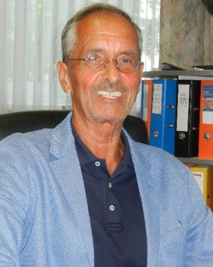 Ein Mann mit Brille und grauen Haaren sitzt in einem Bürostuhl. Er trägt ein hellblaues Sakko über einem dunklen Poloshirt. Im Hintergrund sind farbige Aktenordner sichtbar.