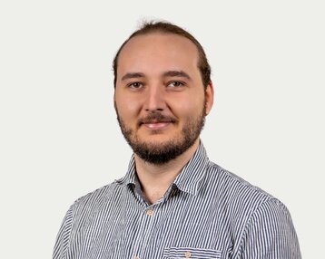 Ein Mann mit Bart und langen Haaren trägt ein gestreiftes Hemd und lächelt in die Kamera vor einem neutralen Hintergrund.