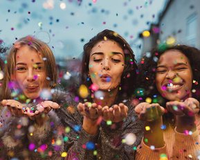 Vier Frauen blasen Konfetti in die Luft, umgeben von bunten Partikel. Sie lächeln und zeigen Freude in einer festlichen Atmosphäre.
