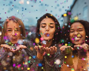 Vier Frauen blasen Konfetti in die Luft, umgeben von bunten Partikel. Sie lächeln und zeigen Freude in einer festlichen Atmosphäre.