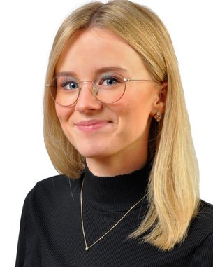 Eine junge Frau mit blonden Haaren und Brille trägt ein schwarzes Oberteil. Sie lächelt leicht und steht vor einem weißen Hintergrund.