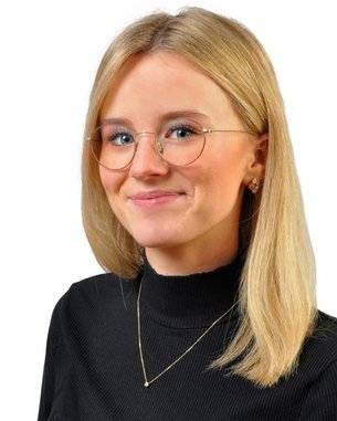 Eine junge Frau mit blonden Haaren und Brille trägt ein schwarzes Oberteil. Sie lächelt leicht und steht vor einem weißen Hintergrund.