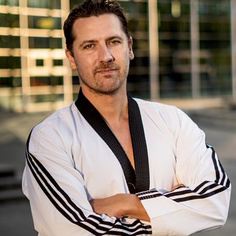 Ein Mann in einem weißen Taekwondo-Anzug mit schwarzem Gürtel steht mit verschränkten Armen. Er hat kurze, dunkle Haare und einen neutralen Gesichtsausdruck. Im Hintergrund sind moderne Gebäude sichtbar.