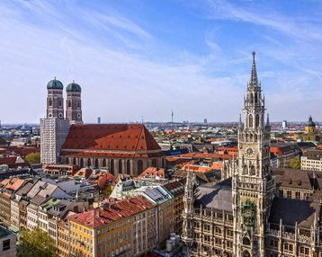 Studienzentrum München | SRH Fernhochschule Moderne Gebäude mit großen Fenstern und einem überdachten Außenbereich. Tische und Stühle unter grünen Sonnenschirmen sind im Vordergrund sichtbar.