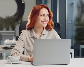 Eine lächelnde Frau mit roten Haaren sitzt an einem Schreibtisch vor einem Laptop. Auf dem Tisch steht eine Tasse Kaffee. Der Hintergrund zeigt eine moderne Büroumgebung.