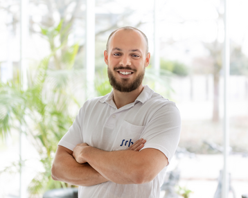 Ein Mann mit Bart und kurzen Haaren steht mit verschränkten Armen in einem hellen Raum. Er trägt ein weißes Hemd mit einem Logo auf der Brust und lächelt freundlich.