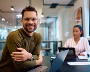 Ein lächelnder Mann mit Brille sitzt an einem Tisch mit einem Laptop, während eine Frau im Hintergrund an einem Tablet arbeitet.