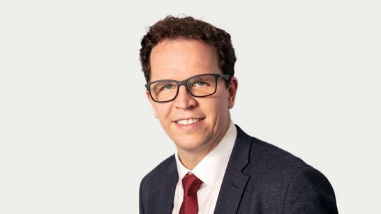 Ein Mann mit Brille und lockigem Haar trägt einen Anzug und eine Krawatte. Er lächelt freundlich in die Kamera.