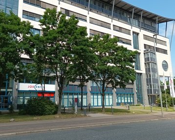 Modernes Bürogebäude mit großen Fenstern, umgeben von Bäumen. Im Vordergrund ist ein Schild mit der Aufschrift "JOB".