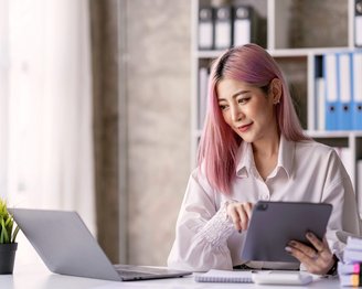 Eine Frau mit pinken Haaren sitzt an einem Schreibtisch, nutzt ein Tablet und schaut auf einen Laptop. Im Hintergrund sind Aktenordner und eine kleine Pflanze sichtbar.