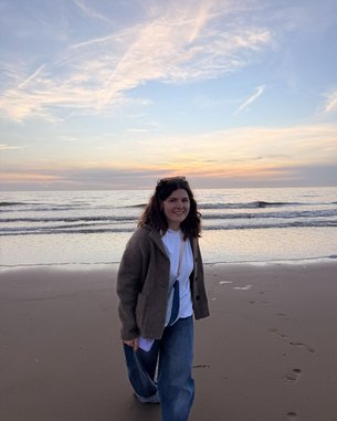 Eine Person mit lockigem Haar trägt einen grauen Pullover und Jeans und geht am Strand entlang, während die Wellen sanft an den Ufer schlagen.