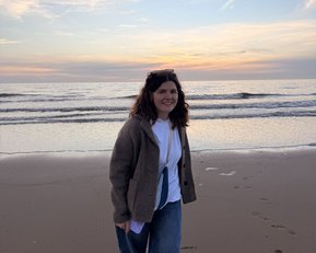 Eine Person mit lockigem Haar trägt einen grauen Pullover und Jeans und geht am Strand entlang, während die Wellen sanft an den Ufer schlagen.