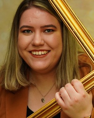 Eine Person mit blonden Haaren hält einen goldenen Bilderrahmen vor ihrem Gesicht. Sie trägt ein braunes Oberteil und lächelt. Der Hintergrund ist unscharf und funkelt in warmen Farben.