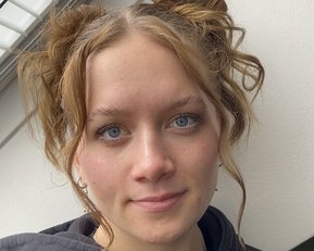 Junge Frau mit blonden, lockigen Haaren, die zu zwei Duttfrisuren gestylt sind. Sie trägt einen schwarzen Hoodie und schaut direkt in die Kamera.