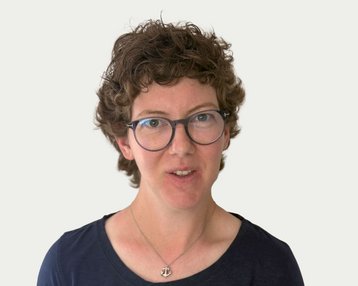 Frau mit lockigem, braunem Haar und Brille, trägt ein schwarzes Oberteil. Sie schaut direkt in die Kamera.