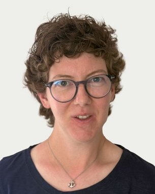 Frau mit lockigem, braunem Haar und Brille, trägt ein schwarzes Oberteil. Sie schaut direkt in die Kamera.