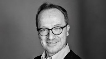 Ein Mann mit Brille und kurzem, hellbraunem Haar lächelt in die Kamera. Er trägt ein Hemd und eine Weste. Der Hintergrund ist unscharf und neutral.