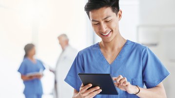 Ein männlicher Gesundheitsdienstleister in blauer Arbeitskleidung nutzt ein Tablet. Im Hintergrund sind zwei weitere Fachkräfte zu sehen, die miteinander sprechen.