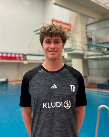 Ein junger Mann steht vor einem Sprungbrett in einem Schwimmbad. Er trägt ein sportliches T-Shirt mit dem Aufdruck "TA" und "KLUDI". Im Hintergrund ist ein Sprungbrett sichtbar.