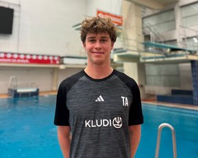 Ein junger Mann steht vor einem Sprungbrett in einem Schwimmbad. Er trägt ein sportliches T-Shirt mit dem Aufdruck "TA" und "KLUDI". Im Hintergrund ist ein Sprungbrett sichtbar.