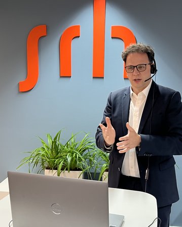 Ein Mann mit Headset steht vor einem Laptop und spricht, während im Hintergrund das Logo "srl" und eine Pflanze sichtbar sind.