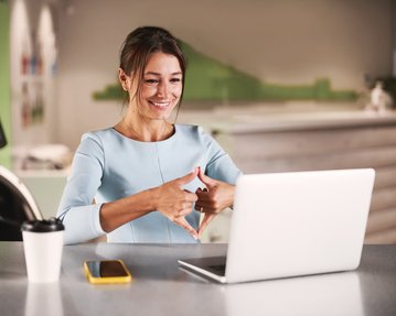 Eine Frau in einem hellblauen Kleid sitzt an einem Tisch und lächelt, während sie mit beiden Händen ein Herzsymbol vor einem Laptop formt.