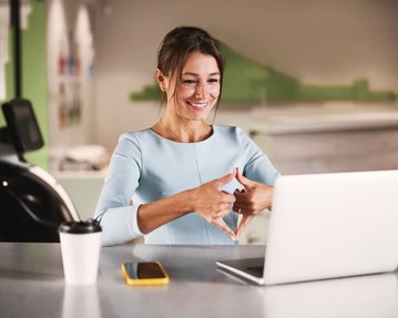 Eine Frau in einem hellblauen Kleid sitzt an einem Tisch und lächelt, während sie mit beiden Händen ein Herzsymbol vor einem Laptop formt.