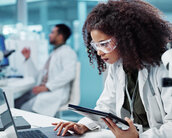 Eine Wissenschaftlerin in einem Labor arbeitet konzentriert an einem Laptop und hält ein Tablet in der Hand. Im Hintergrund ist ein Kollege zu sehen.