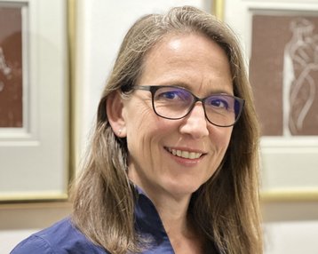 Eine Frau mit langen, glatten Haaren und einer blauen Bluse steht lächelnd vor gerahmten Kunstwerken. Sie trägt eine Brille und hat einen freundlichen Ausdruck.