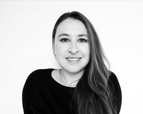 Eine Frau mit langen, braunen Haaren und einem freundlichen Lächeln trägt ein schwarzes Oberteil. Der Hintergrund ist neutral und hell.