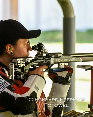 Eine Schützin in Sportkleidung zielt mit einem Luftgewehr. Sie trägt Ohrstöpsel und hat ihre Haare zu einem Zopf gebunden. Der Hintergrund zeigt eine Schießanlage.