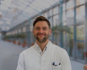 Ein Mann in einem weißen Kittel steht lächelnd in einem modernen, hellen Raum mit Pflanzen im Hintergrund.