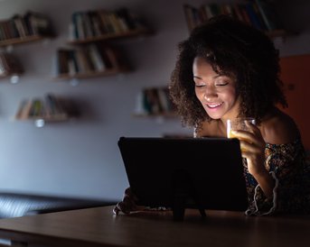 Eine Frau mit lockigem Haar sitzt an einem Tisch und schaut auf ein Tablet, während sie ein Getränk in der Hand hält. Im Hintergrund sind Bücherregale sichtbar.