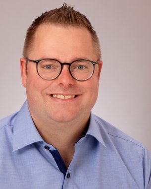 Ein lächelnder Mann mit Brille trägt ein hellblaues Hemd. Der Hintergrund ist neutral und hell.