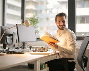 Ein Mann sitzt an einem Schreibtisch in einem modernen Büro, hält ein Buch in der Hand und lächelt. Auf dem Tisch befinden sich Computerbildschirme und ein Kopfhörer.