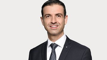 Ein Mann in einem Anzug mit Krawatte lächelt in die Kamera. Er hat kurze, dunkle Haare und trägt ein weißes Hemd unter einem dunklen Sakko. Der Hintergrund ist neutral und hell.