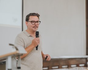 Ein Mann mit Brille hält ein Mikrofon und spricht vor einer Präsentation in einem Seminarraum. Er trägt ein helles, kurzärmliges Hemd und hat einen gepflegten Schnurrbart.