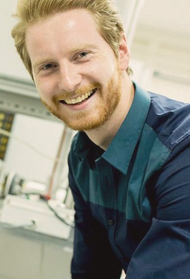 Ein lächelnder Mann mit blonden Haaren trägt ein blaues Hemd und arbeitet an einer technischen Apparatur in einem Labor. Kabel und Geräte sind im Hintergrund sichtbar.