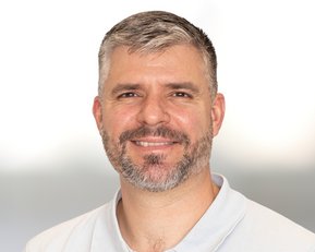 Ein Mann mit grauen Haaren und Bart lächelt freundlich. Er trägt ein helles Polohemd und hat einen neutralen Hintergrund.