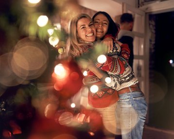 Zwei Frauen umarmen sich lächelnd, während sie festliche Pullover tragen. Im Hintergrund sind verschwommene Lichter und Weihnachtsdekoration zu sehen.
