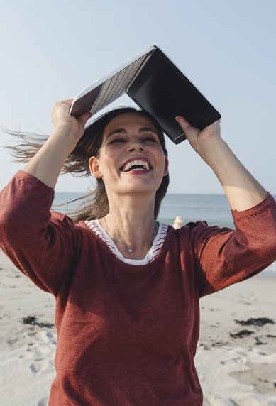 Frau mit einem Buch über dem Kopf, lächelnd am Strand, mit Meer im Hintergrund und leichtem Wind, der ihre Haare bewegt.
