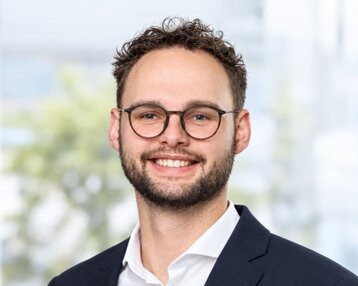 Ein junger Mann mit Brille und Bart lächelt, trägt einen Anzug und ein Hemd. Der Hintergrund zeigt unscharfe, helle Farben.